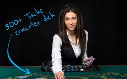 Ein Live Dealer sitzt an einem Blackjack Spieltisch. Sie blickt direkt in die Kamera. In ihrer linken Hand hält sie einige Spielkarten. Mit der rechten Hand langt sie über die Spielchips hinweg auf das Setztableau.