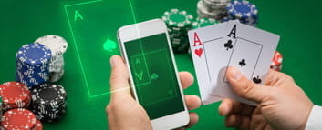 Zwei Hände vor einem Pokertisch mit Chips. In der rechten Hand befinden sich zwei Asse und in der linken Hand ein Smartphone.