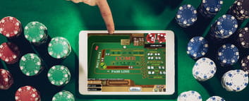 Ein Finger zeigt auf ein Tablet mit dem Online Spiel Craps, welches in der Mitte liegt. Links und rechts davon befinden sich Casino Chips.