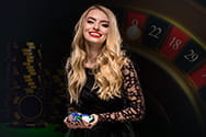 Eine Dame hält Casino Chips in der Hand und im Hintergrund ist ein Roulettekessel zu sehen.