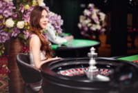 Ein Crouper sitzt in einem blumengeschmückten Raum schräg links hinter dem Roulettekessel und lächelt in die Kamera. Es handelt sich um einen Live Casino Spieltisch, der von Evolution Gaming betrieben wird.