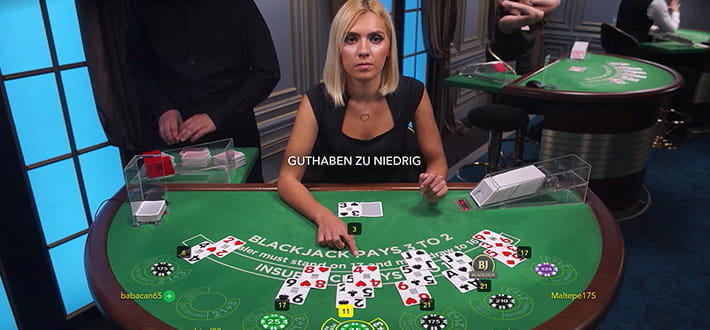 Live Blackjack H Spieltisch, bereitgestellt von Evolution Gaming im Live Bereich von 777 Casino. Der Dealer fragt den ihm direkt gegenüber sitzenden Spieler, ob er eine weitere Spielkarte erhalten möchte. Ein weiterer Dealer mischt leicht links versetzt vom Spieltisch neue Karten. Rechts hinten sind weitere Spieltische zu sehen.