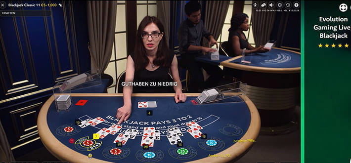 Live Blackjack Classic von Evolution Gaming im Live Casino von Casino Room. Der Dealer zeigt mit der Hand auf die Karten des Spielers, der als nächstes entscheiden muss, ob er eine weitere Karte ziehen möchte. Im Hintergrund befindet sich ein weiterer Spieltisch.