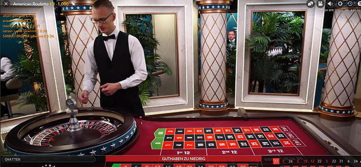 Live American Roulette von Evolution Gaming im Live Casino von DrückGlück. Der Croupier hat gerade die Kugel in den Kessel geworfen. Im Hintergrund sind säulenartige Dekorationselemente sowie weitere Spieltische zu erkennen.
