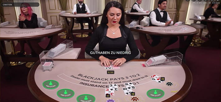 Ein Blackjack L Live Spieltisch im Live Casino, bereitgestellt von Evolution Gaming. Der Dealer deckt gerade seine zweite Karte auf. Im Hintergrund sieht man weitere Live Spieltische.
