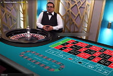 Auf diesem Bild sieht man einen männlichen Live Dealer vor einem Roulettekessel. Vor ihm ist der Setztisch.