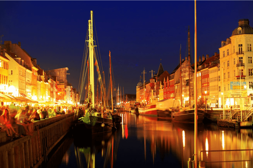 Auf dem Bild ist das Hafenbecken Nyhavn in der dänischen Hauptstadt Kopenhagen zu sehen. Das Foto ist bei Nacht aufgenommen und die alten Straßenlaternen beleuchten die Segelschiffe, bunten Fassaden in den Straßen.