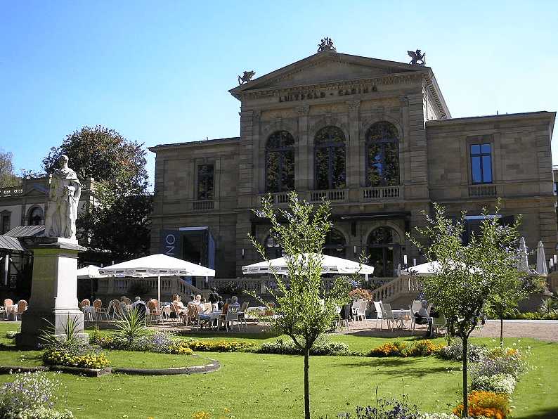 Das Bild zeigt die Aussenansicht des Casinos in Bad Kissingen. Zu sehen ist eine zweistöckige Villa und eine Gartenlage mit einer Statue.