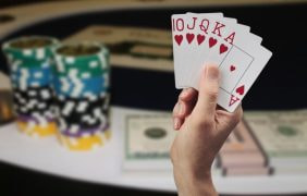 Ein Spieler hält ein Royal Flush in Herzkarten in der Hand vor einem Poker-Tisch, auf dem auch Chips gestapelt sind.