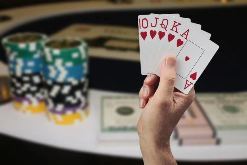 Ein Spieler hält ein Royal Flush in Herzkarten in der Hand vor einem Poker-Tisch, auf dem auch Chips gestapelt sind.