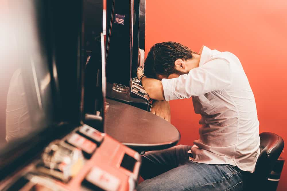 Ein Mann sitzt deprimiert an einem Spielautomaten im Casino