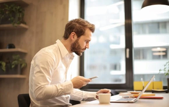 Ein Mann im weißen Hemd schaut auf sein Smartphone und seinen Laptop