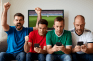 Vier Männer sitzen auf einer Couch und schauen auf ihre Smartphones. Einige jubeln, andere sind zerknirscht.