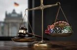 Eine goldene Waage mit Richterhammer auf der einen und Pokerchips sowie Rouletteelementen auf der anderen Seite steht in einem dunklen Raum. Im Hintergrund ist unscharf ein deutsches Regierungsgebäude zu sehen.