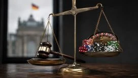 Eine goldene Waage mit Richterhammer auf der einen und Pokerchips sowie Rouletteelementen auf der anderen Seite steht in einem dunklen Raum. Im Hintergrund ist unscharf ein deutsches Regierungsgebäude zu sehen.