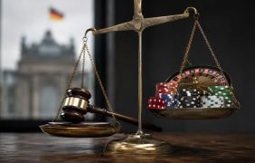 Eine goldene Waage mit Richterhammer auf der einen und Pokerchips sowie Rouletteelementen auf der anderen Seite steht in einem dunklen Raum. Im Hintergrund ist unscharf ein deutsches Regierungsgebäude zu sehen.