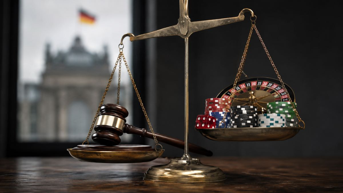 Eine goldene Waage mit Richterhammer auf der einen und Pokerchips sowie Rouletteelementen auf der anderen Seite steht in einem dunklen Raum. Im Hintergrund ist unscharf ein deutsches Regierungsgebäude zu sehen.