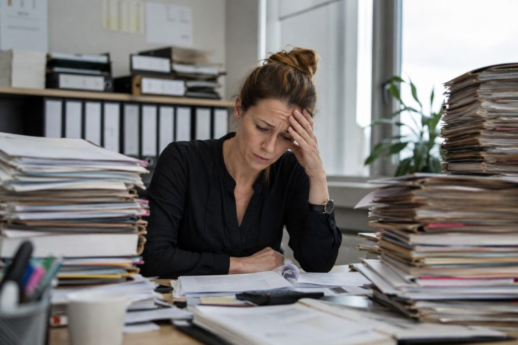 Ein Büro in einer Behörde im Saarland. Ein Berg voller Akten und eine Mitarbeiterin sieht überfordert aus.